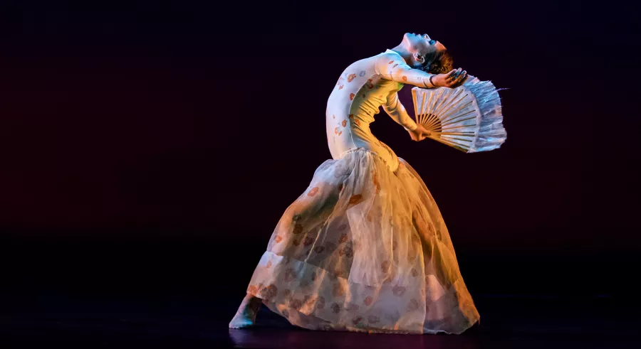 A side profile of a dancer on a dark stage shows them bending backward with one leg forward and the other back, holding a handheld fan with their arms reaching out behind, and wearing a white body suit and loose, transparent pants.