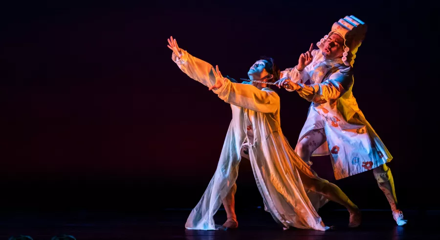 Two dancers stand on a dark stage dressed in white with pops of color. One dancer stands slightly behind the other with an object held at the other dancer's neck, who gazes upward with their arms in an outstretched position.