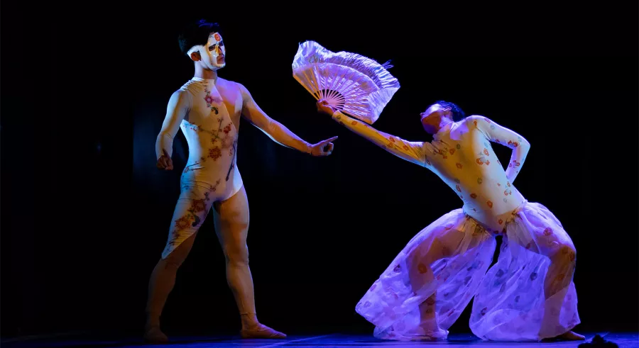 A stage lit in blue shows two dancers in white clothing facing one another. One dancer stands with their legs bent at the knee facing outward to their sides, outstretching their hand and holding a fan toward the other dancer, who stands straight with their arms slightly out and forward.