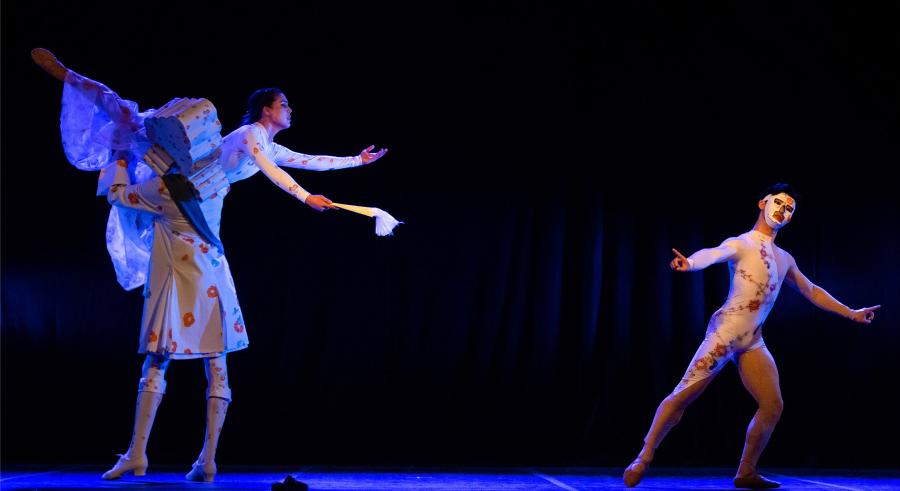 A dark stage lit in blue shows three dancers in white clothing. One dancer holds another up in a lift position, who reaches out and down toward the third dancer, who stands a few feet away, leaning slightly right with their arms outstretched forward.