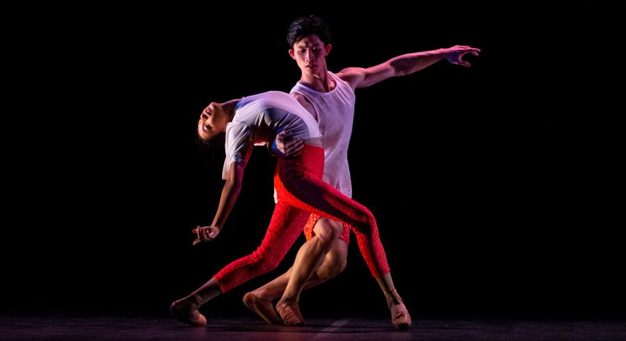 Two dancers dressed in white and red stand on a dark stage. One holds the other, bent backward at the waist with one leg pointed forward with a slight bend at the knee.