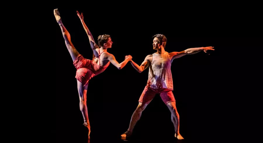 A dark stage shows two dancers facing one another and holding hands with a slight bend at their elbows. The dancer on the left stands with one leg on the ground and the other lifted directly up and aligned with the other arm. The dancer on the right stands firmly with both feet on the ground and one arm stretched parallel to the floor.