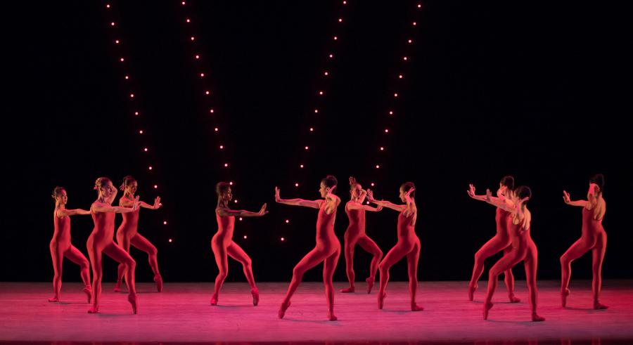 A stage lit in pink shows a group of dancers dressed in red standing facing both away and toward one another with one leg planted on the ground and the other slightly lifted with a bend at the knee.