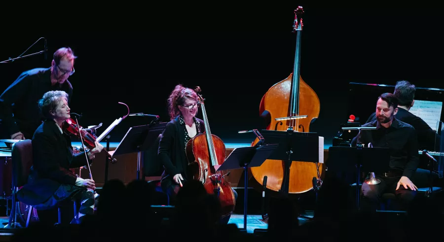 Six musicians dressed in black perform onstage with music stands in front of them. The camera focuses on a drum player, violinist, cellist, and horn player, with a pianist and bass positioned slightly behind them.