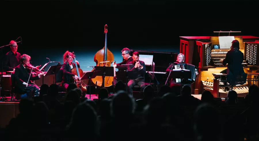 An ensemble of seven musicians including a drum player, violinist, bassist, cellist, horn player, pianist, accordionist, and pipe organist dressed in black perform onstage with a dark background and pink lighting. 