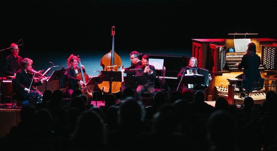 An ensemble of seven musicians including a drum player, violinist, bassist, cellist, horn player, pianist, accordionist, and pipe organist dressed in black perform onstage with a dark background and pink lighting. 