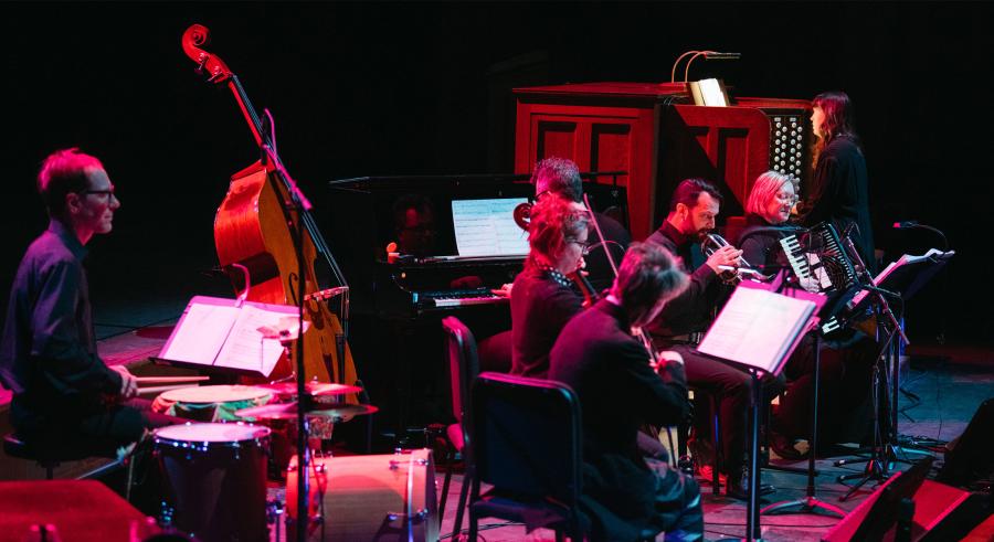 An ensemble of seven musicians dressed in black perform onstage with a dark background and pink lighting. A drummer sits closest to the camera, with the other musicians' backs and sides towards the lens.