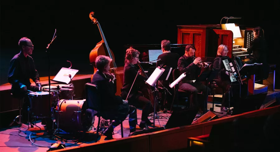 An ensemble of seven musicians including a drum player, violinist, bassist, cellist, horn player, pianist, accordionist, and pipe organist dressed in black perform onstage with a dark background and pink lighting. 