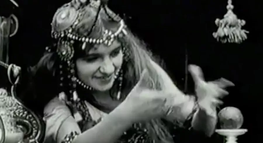 A black and white image shows a person dressed in jeweled headwear sitting at a table focused on an object in front of them. Their hands extend toward the object.