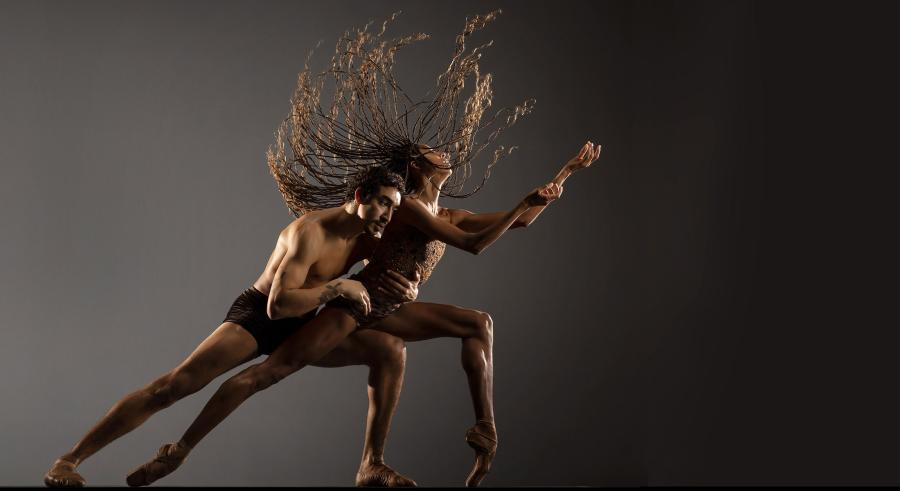 Two dancers appear in front of a gray backdrop facing the right of the frame with one leg outstretched behind them and the other bent at the knee perpendicular to the floor. One dancer's long hair flips up through the air while the other dancer leans on the back of their shoulder.