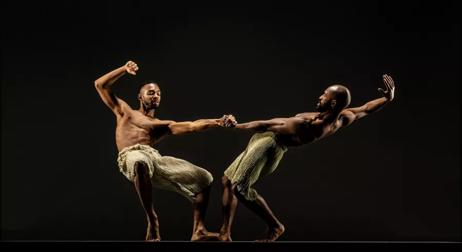 Two dancers appear on a dark stage holding each other by the hand while they outstretch their bodies way from one another.