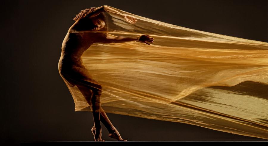 A dancer appears within a stretched, gold fabric, molded to the shape. One arm is lifted above their head, with the other stretched out parallel to the floor. Their feet are pointed, with one knee slightly bent and the other leg stretched out.