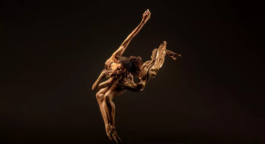 A dancer dressed in gold appears on a dark stage bending forward toward the camera, their feet pointed and crossed and an arm extended up and back. A piece of their clothing flows up in the air along their arm.