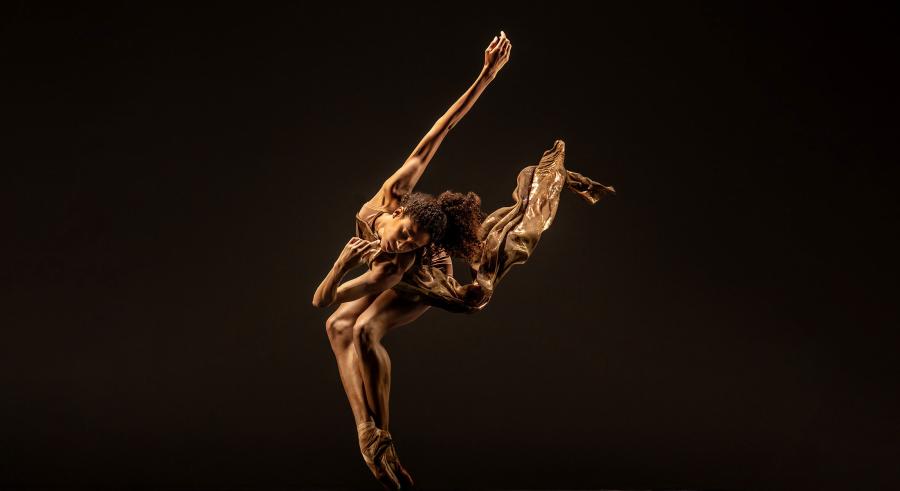 A dancer dressed in gold appears on a dark stage bending forward toward the camera, their feet pointed and crossed and an arm extended up and back. A piece of their clothing flows up in the air along their arm.