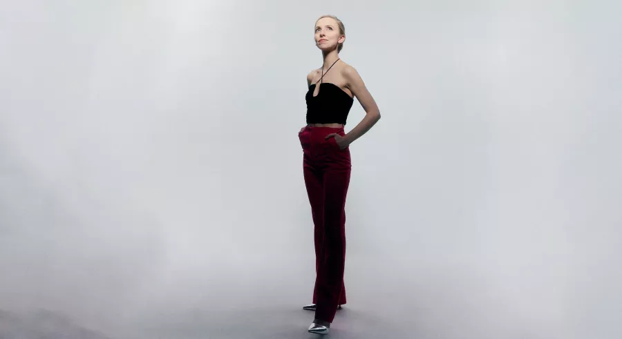A person with blond hair dressed in a black top and red pants stands in front of a light gray backdrop, looking up and off into the distance.