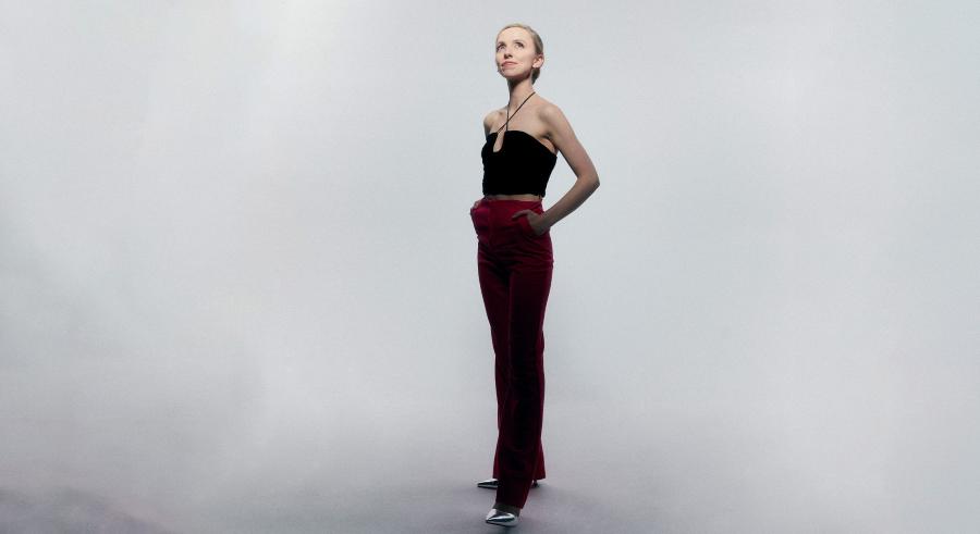 A person with blond hair dressed in a black top and red pants stands in front of a light gray backdrop, looking up and off into the distance.