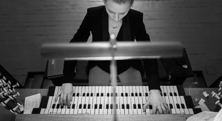 A black and white image shot from above shows a person wearing a blazer looking down and playing a pipe organ. 