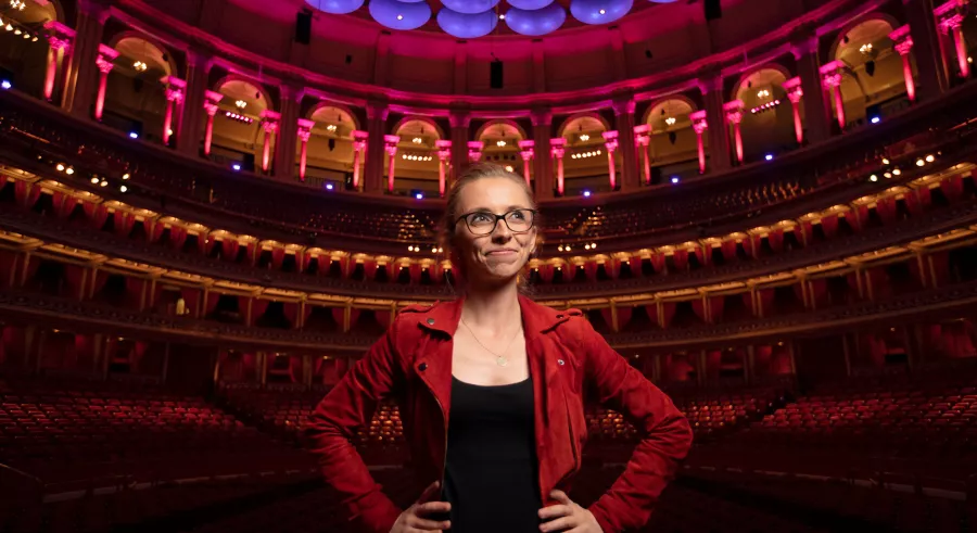A person with blond hair wearing a red jacket, black top, and glasses stands in a theater lit in pink, red, and blue. They hold their hands on their hips and look off into the distance.