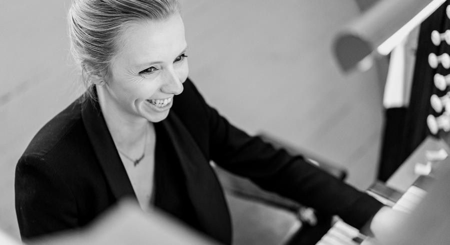 A black and white image focuses on a person with light hair smiling in a black blazer while playing a pipe organ.