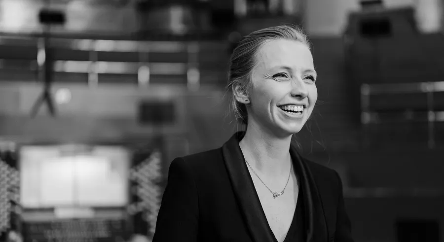 A black and white portrait shows a person with light hair wearing a black blazer, holding a smile and looking off into the distance. A blurred pipe organ appears behind them.