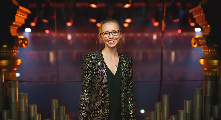 A person with blond hair wearing glasses and a shiny blazer stands in front of the pipes of an organ with their arms to their sides. 