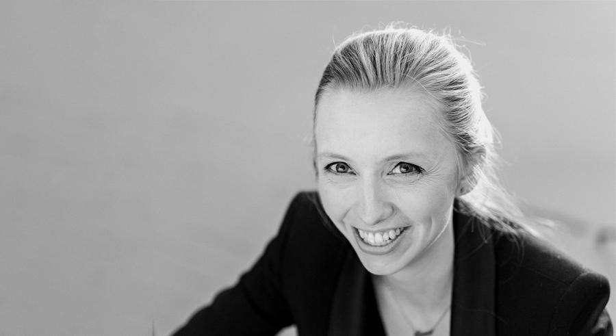 A black and white portrait shows a person with light hair wearing a black blazer, smiling at the camera.