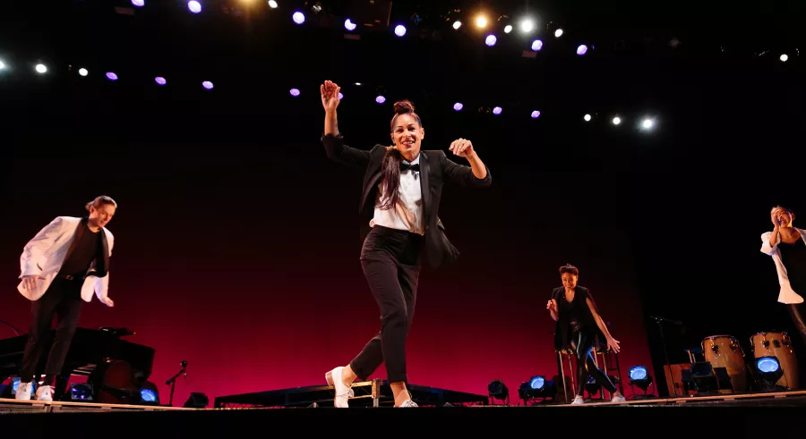 Four dancers are onstage dressed in tap shoes with black pants and a mix of black and white tops. One dancer is slightly downstage, while the other Three dancers form a semicircle around them.