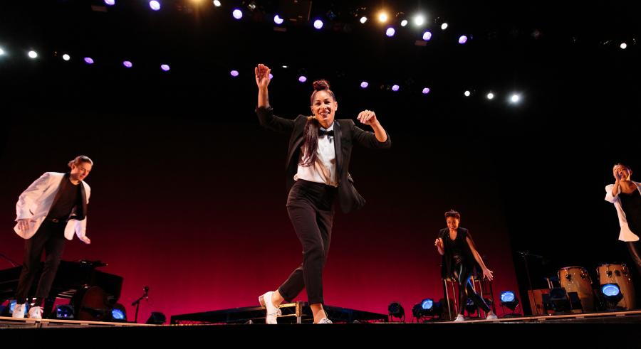 Four dancers are onstage dressed in tap shoes with black pants and a mix of black and white tops. One dancer is slightly downstage, while the other Three dancers form a semicircle around them.