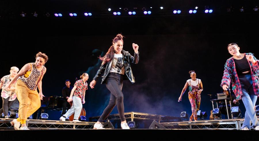 Five dancers and one musician appear in-motion onstage in casual clothing. Three dancers are slightly downstage, while the other three dancers and one musician are somewhat behind them on a raised platform.