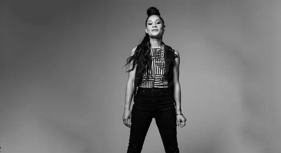 A black and white portrait of a person standing directly toward the camera with her hair half up in a bun wearing dark pants and a vest over a striped tank top.