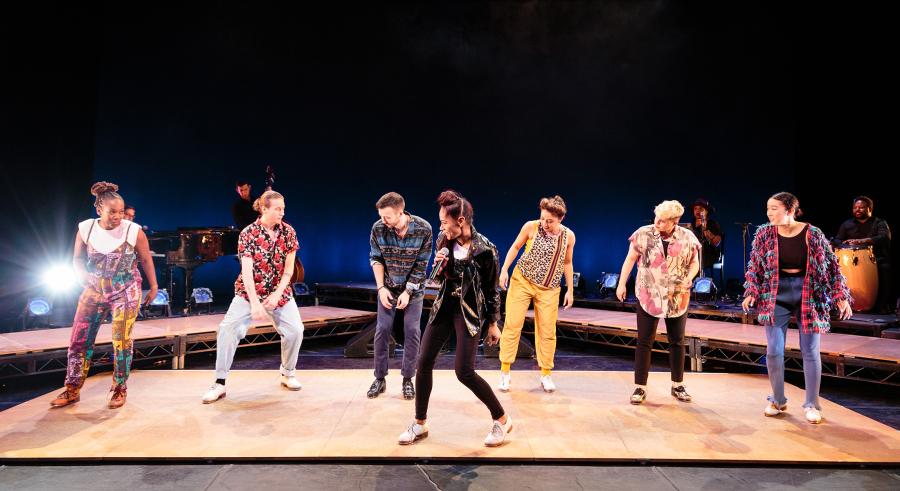 Six dancers stand on stage wearing casual clothing and tap shoes. One dancer is slightly downstage while the other five dancers form a line behind them.