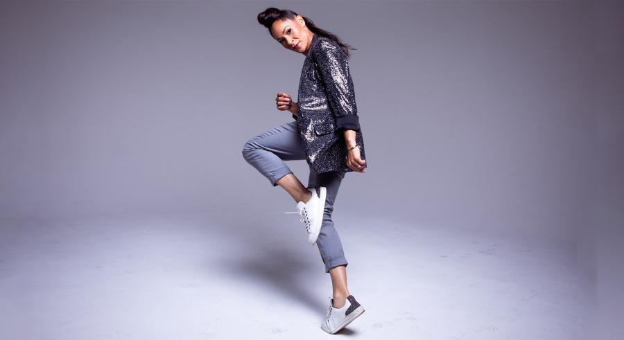 A dancer dressed in grey pants, a shiny blazer, and sneakers balances on one foot in a running position and looks at the camera.