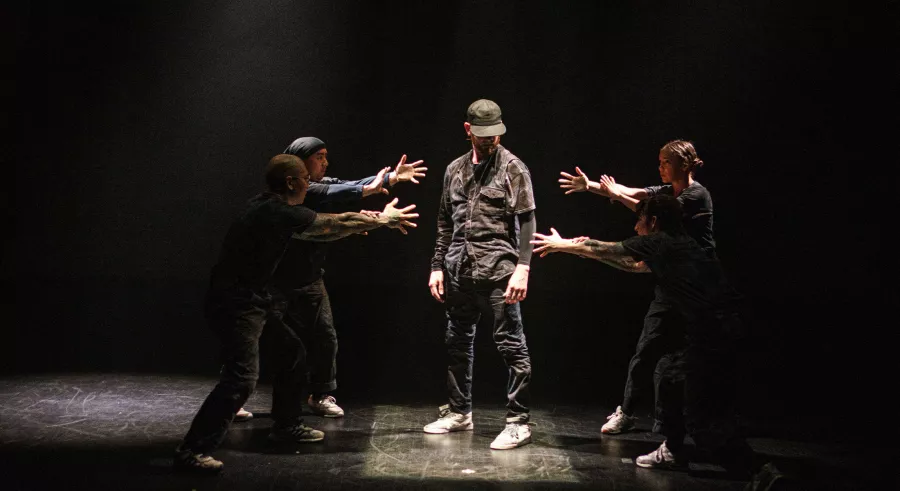 A dancer dressed in dark clothing, a cap, and white sneakers stands in the middle of a spotlight on a dark stage. Four additional dancers stand around them with their arms outstretched pointing toward the middle dancer.