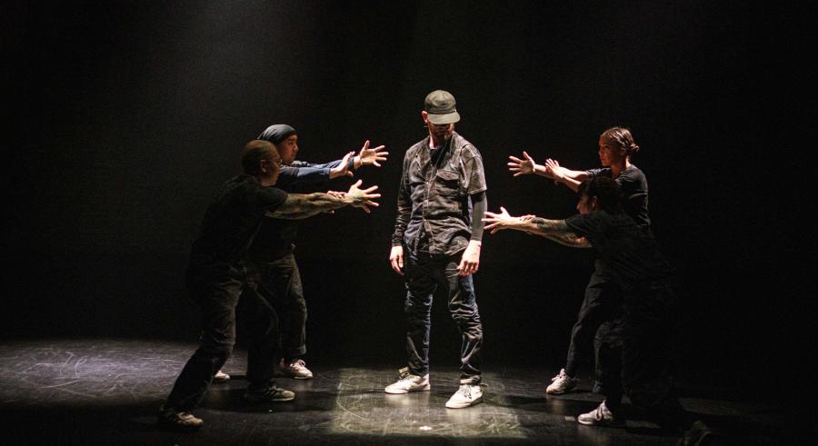 A dancer dressed in dark clothing, a cap, and white sneakers stands in the middle of a spotlight on a dark stage. Four additional dancers stand around them with their arms outstretched pointing toward the middle dancer.