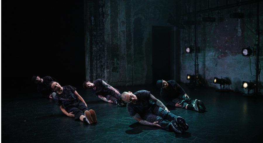 Four dancers dressed in casual dark clothing and sneakers sit on a dark stage with their legs oustretched forward, feet pointing straight up, and slightly leaning on one hip with their bodies pointed to the left. An industrial, concrete wall is slightly visible around them.