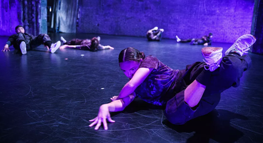 A group of dancers lay in various positions across a stage lit in purple. One dancer sits in the forefront, one leg crossed over the other, pointing up and slightly curled. They rest their head on the shoulder of one arm planted on the ground and fall over the other arm, outstretched forward with a bend in the wrist and fingers spread wide.