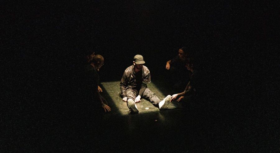 A dancer dressed in dark clothing, a cap, and white sneakers sits in the middle of a spotlight on a dark stage. Additional dancers sit and kneel around them in the darkness that surrounds the area lit by the spotlight.