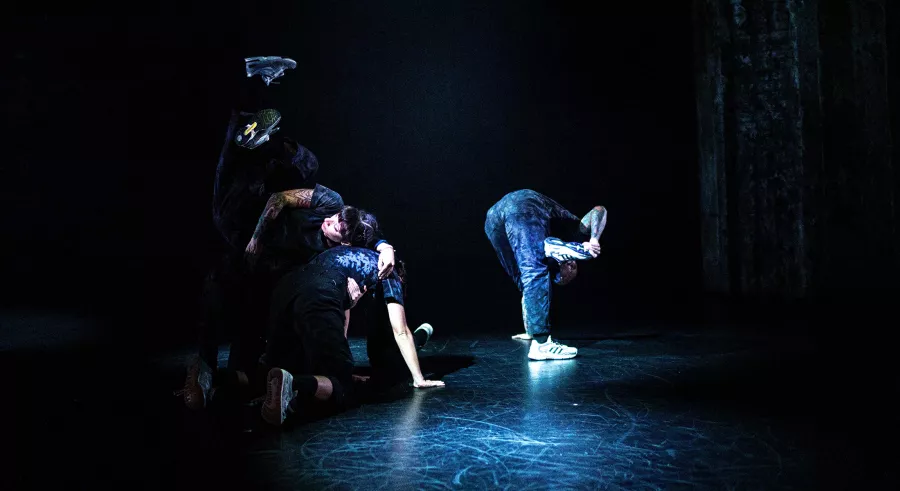 A group of dancers are in various positions across a stage lit in blue. Their bodies are bent forward as the grab their feet with one hand and the other is on the ground.