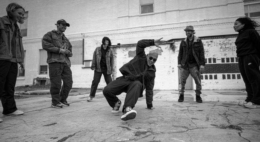 A black and white image shows five people formed in a semi-circle around a dancer dressed in pants and a jacket with sneakers who is crouching with one leg forward and slightly bent and the other planted behind them. They hold one arm above their head, with the remaining arm stable on the ground.