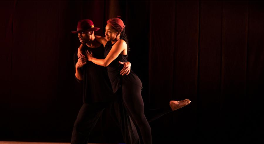 Two dancers in black clothing and maroon headwear dance onstage with their arms wrapped around one another, looking to the left. The dancer on the right one leg lifted parallel to the floor while the other has both feet planted.
