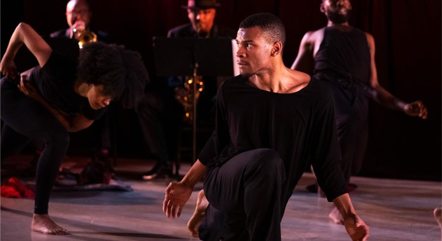 Three dancers and two musicians perform onstage wearing all black. 1 dancer stands slightly downstage, kneeling and looking to the left with the other two slightly behind in various positions.