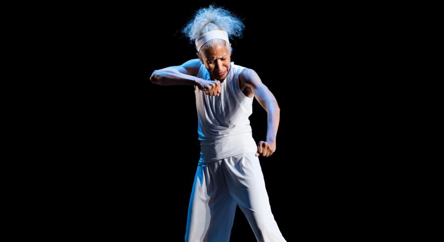 A gray-haired dancer wearing a loose-fitted white tank top and pants stands onstage towards the camera, looking down with one arm raised parallel to the floor and the other slightly lower, their hands clenched.