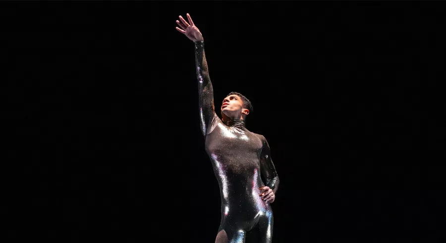 A dancer dressed in a shiny, fitted jumpsuit poses looking up with one arm stretched toward the ceiling and the other placed on their hip against a black background.