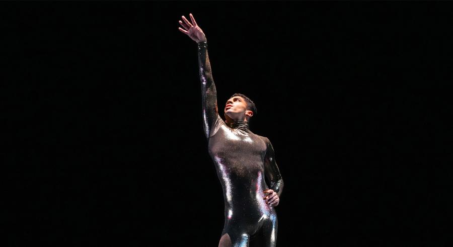 A dancer dressed in a shiny, fitted jumpsuit poses looking up with one arm stretched toward the ceiling and the other placed on their hip against a black background.