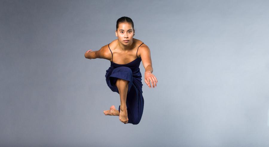 A dancer dressed in blue appears in front of a gray backdrop, jumping in the air and facing directly at the camera.