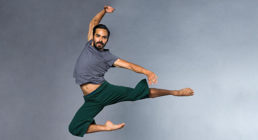 A dancer dressed in a gray top and green pants appears in front of a gray backdrop, leaping in the air with both legs pointing to the left, one extended straight and the other bent at the knee. They hold one arm above them, and the other stretched out to the left.