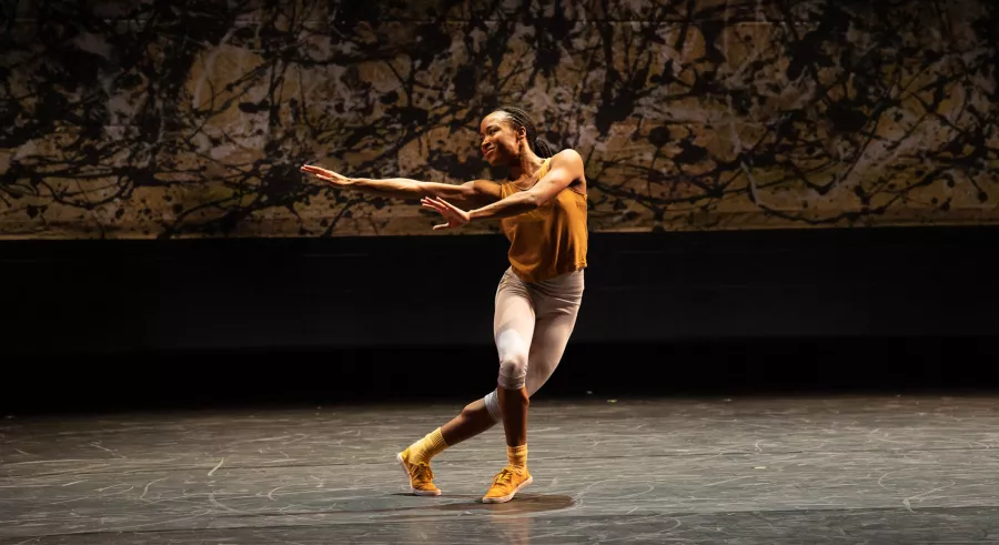 A dancer performs onstage dressed in a yellow top and shoes, and white pants. They stretch their arms out to the left and across their body, which faces the camera. One leg stands in front of the other, slightly bent with the other crossed behind.