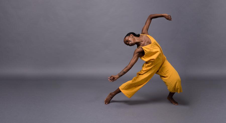 A dancer wearing a yellow, sleeveless jumpsuit poses against a gray backdrop. They face the camera with their head bent to the left and parallel to the ground with one leg stretched out to the left and the other bent slightly at the knee. One arm drapes across the outstretched leg, the other raised up and bent at the elbow.
