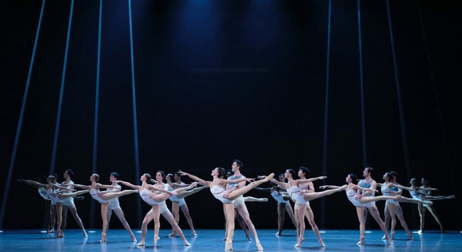 A group of dancers stand onstage in a line, partnered up, and looking to the left. The dancers in front hold one leg on the ground and the other pointed out behind them, while the other dancers hold their waists. They wear matching white, fitted clothing.