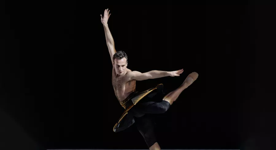 A dancer wearing black leggings and a structured tutu-like piece poses on a dark stage with their arms at a ninety-degree angle, looking down with one leg planted and the other bent at the knee and pointing behind them.
